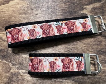 May include: Two black keychains with a white and pink floral pattern featuring highland cows. The keychains have silver key rings.