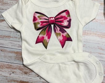 Könnte beinhalten: Ein weißer kurzärmeliger Babybody mit einer großen, mittig platzierten Schleifengrafik. Die Schleife zeigt ein camouflage-inspiriertes Muster in den Farben Pink, Dunkellila und Olivgrün. Das Kleidungsstück hat einen Rundhalsausschnitt und Druckknöpfe.