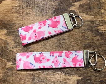 May include: Two pink pig keychains with silver key rings. The keychains are made of fabric with a white background and pink pig illustrations.