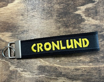 Könnte beinhalten: Schwarzer Stoff-Schlüsselanhänger mit dem Namen "CRONLUND" in Gelb gestickt. Der Schlüsselanhänger hat einen silberfarbenen Metallverschluss und Schlüsselring. Der Hintergrund ist eine Holzoberfläche.