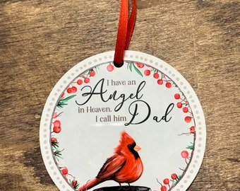 May include: A white round ornament with a red cardinal perched on a wooden stump. The ornament has a red ribbon attached and says "I have an Angel in Heaven. I call him Dad."