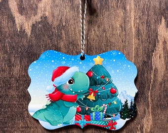 Könnte beinhalten: Ein Weihnachtsbaumschmuck mit einem grünen Dinosaurier, der einen roten Weihnachtsmann-Hut und einen Schal trägt, neben einem geschmückten Weihnachtsbaum mit Geschenken und einem Stern obenauf. Der Schmuck ist auf einem Holzgrund.