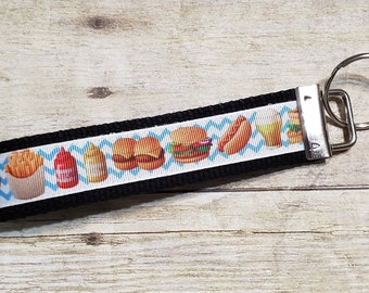 Könnte beinhalten: Schwarzer Stoff-Schlüsselanhänger mit einem weißen Band mit einem sich wiederholenden Muster von Fast-Food-Artikeln wie Burgern, Pommes Frites und Hotdogs. Das Band hat ein blaues Zickzack-Design. Ein silberner Metall-Schlüsselring ist befestigt.