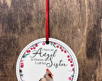 May include: A round, white ornament with a red ribbon for hanging. The ornament features a cardinal bird illustration and the text "I have an Angel in Heaven. I call her Sister" surrounded by a wreath of red berries and green leaves.