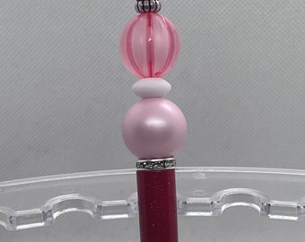 May include: A pink and burgundy pen with a pink and white bead design. The pen has a sparkly burgundy finish and a silver band around the middle.