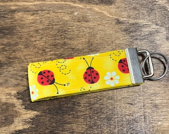May include: A yellow fabric keychain with a ladybug pattern and a silver key ring. The fabric has white daisies and black dotted lines.