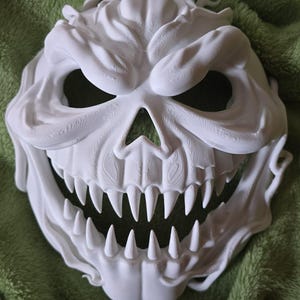 May include: A white, 3D-printed mask with a menacing skull design. The mask features sharp teeth, deep eye sockets, and a textured surface, creating a spooky and detailed appearance. The mask is likely for Halloween or cosplay.