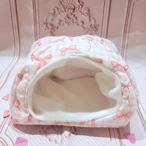May include: A pink and white pet bed with a bow pattern. The bed has a soft, white interior and a gingham exterior with pink bows. The bed is designed with an open entrance and is likely for a small animal.