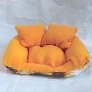 Peut inclure: Un petit lit pour animaux de compagnie avec trois coussins orange. Le lit est jaune vif avec un motif de tournesol autour de la base. Le lit est fait d'une matière douce et moelleuse.