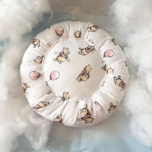 May include: A round, white baby pillow featuring a repeating pattern of Winnie the Pooh characters and pink balloons. The pillow has a ruffled edge and is set against a soft, cloudy background.