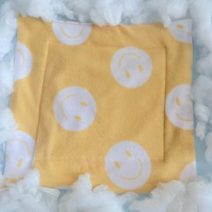 May include: Yellow fleece pet bed with white smiley face pattern. The bed is square with a stitched border. The bed is surrounded by fluffy white clouds. The bed is designed for small pets.