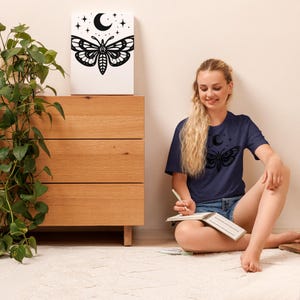 Könnte beinhalten: Ein marineblaues T-Shirt mit einem schwarzen Motten-Design, einem Mond und Sternen. Ein weißer Leinwanddruck auf einer Holzkommode zeigt ein ähnliches Motten-Design. Der Raum hat eine neutrale Farbpalette.