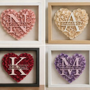 Op de afbeelding: Vier ingelijste bloemstukken in de vorm van een hart. Elk hart is gevuld met papieren bloemen en heeft een gepersonaliseerde naam en initiaal. De frames zijn wit, zwart en van natuurlijk hout, met roze, crème, rode en paarse bloemen.