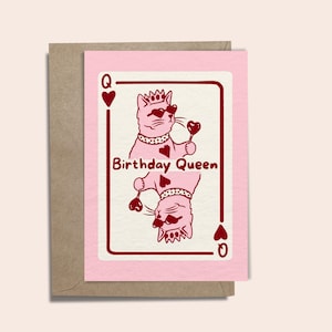 May include: A pink greeting card with a playing card design, featuring a cat illustration. The cat wears sunglasses and a crown, holding a heart-shaped lollipop. The text "Birthday Queen" is displayed in the centre of the card.