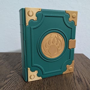 May include: A teal-colored rectangular box designed to resemble a book, featuring gold-toned corner accents and a central circular emblem displaying a raised paw print design. The exterior has a textured finish with molded borders and a side latch.