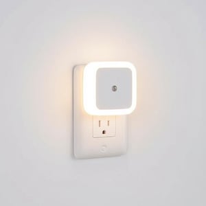 Op de afbeelding: Een wit nachtlampje dat is aangesloten op een wit stopcontact aan een witte muur. Het nachtlampje heeft een vierkant, gloeiend oppervlak met een kleine, cirkelvormige sensor. Het stopcontact heeft twee sleuven en een aardingsgat.
