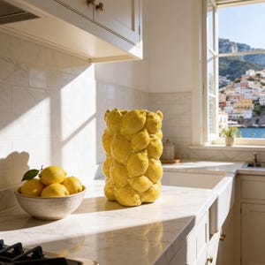 May include: A bright yellow ceramic vase with a textured, bulbous design sits on a marble countertop next to a bowl of lemons. The kitchen setting features white cabinets, a gas stove, and a window overlooking a coastal town.