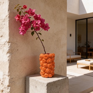 May include: An orange ceramic vase with a textured, bubble-like surface holds a vibrant pink flowering branch. The vase sits on a concrete block, with a neutral-toned wall in the background. The scene evokes a sense of modern design and natural beauty.