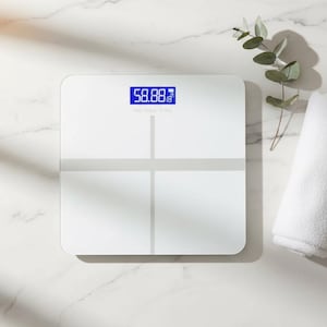 Peut inclure: Balance de salle de bain numérique blanche avec un écran bleu affichant 26,66 kg. La balance a un design en croix et est sur une surface en marbre. Une branche de verdure et une serviette blanche sont à côté.