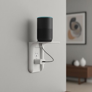 May include: A black smart speaker with a blue accent ring sits on a white shelf attached to a wall outlet. A white charger is plugged into the outlet, with a black cable connected to the speaker.