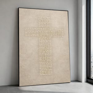 Puede incluir: Una impresión artística en lienzo beige enmarcada con un diseño en forma de cruz lleno de texto religioso en una fuente gris claro. La obra de arte se exhibe contra una pared blanca, con un marco negro.