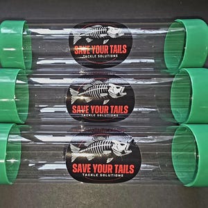 Può includere: Tre tubi di plastica trasparente con tappi verdi. Ogni tubo ha un adesivo nero e rosso con una grafica di scheletro di pesce e il testo "SAVE YOUR TAILS TACKLE SOLUTIONS". I tubi sono disposti orizzontalmente.