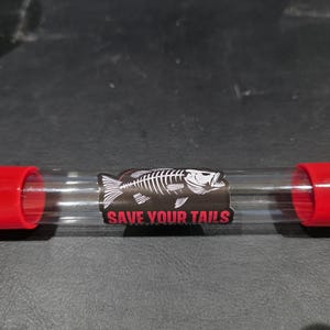 May include: A clear plastic tube with red caps on each end. A black sticker with a white fish skeleton and the words "SAVE YOUR TAILS" is in the center of the tube.