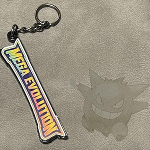 May include: A keychain with a rectangular charm displaying "MEGA EVOLUTION" in a rainbow gradient. The keychain has a silver ring and chain. A grey image of a cartoon character is next to the keychain.