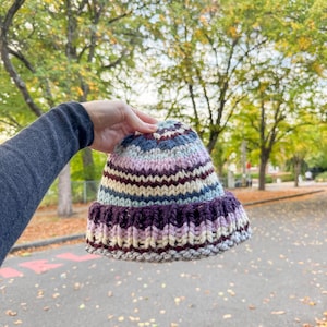 May include: A chunky knit beanie featuring horizontal stripes in shades of deep purple, light pink, cream, and slate blue. The hat has a thick, ribbed folded brim and a rounded crown, constructed from thick yarn with a visible, textured stitch pattern.