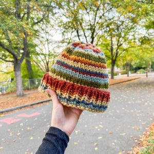 May include: A hand-knit beanie featuring a chunky, ribbed texture and a multi-colored striped pattern. The hat displays horizontal bands of deep red, olive green, mustard yellow, and light blue yarn, finished with a thick, folded brim.