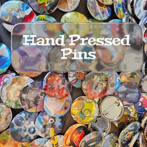 May include: A collection of colorful, hand-pressed pins with various designs. The pins feature images of cartoon characters and other graphics. The text "Hand Pressed Pins" is displayed on a silver background.