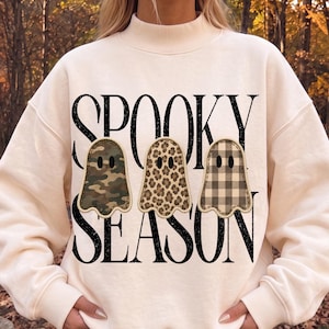 May include: A cream-coloured long-sleeve sweatshirt featuring the black text SPOOKY SEASON. Below the text are three ghost-shaped patches with stitched edges, displaying a green camouflage, a brown leopard print, and a tan plaid pattern.