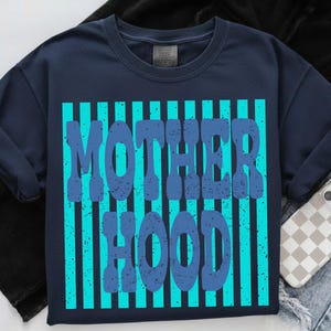 May include: A navy blue t-shirt featuring a graphic design with vertical bright turquoise stripes. The text MOTHER HOOD is printed in a distressed, dark blue, bold font over the stripes. The shirt has a crew neckline and short sleeves.