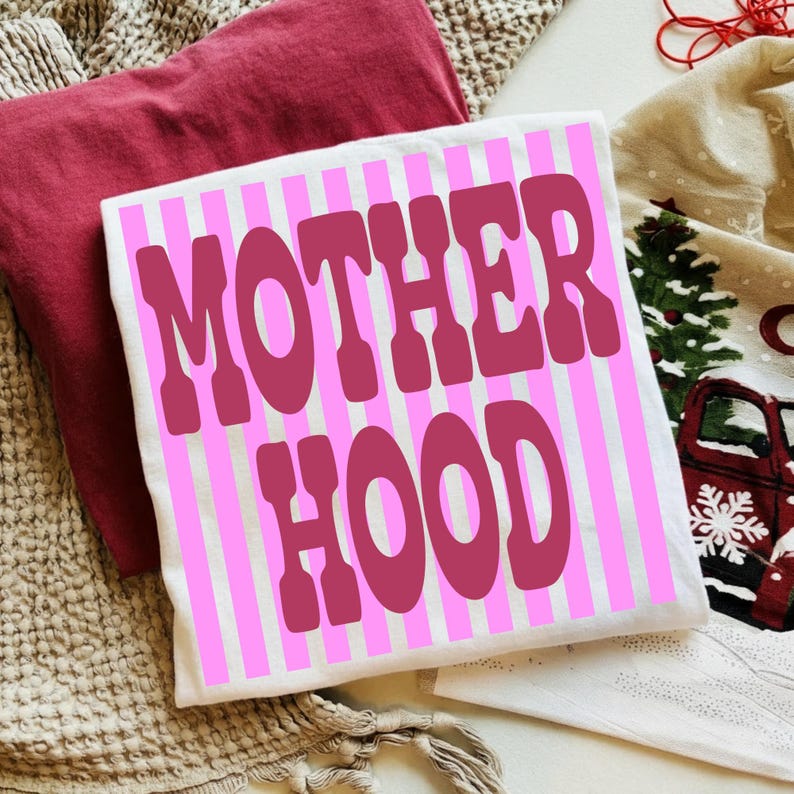 May include: A white t-shirt with pink vertical stripes and the word "MOTHERHOOD" in a bold, retro font. The shirt is displayed on a textured surface with a maroon pillow and a festive holiday-themed towel in the background.