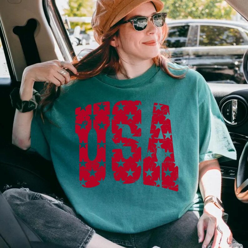 May include: Teal-colored t-shirt with the word "USA" in large, distressed red letters filled with star patterns. The shirt has short sleeves and a relaxed fit. The person in the image is wearing sunglasses and a hat.