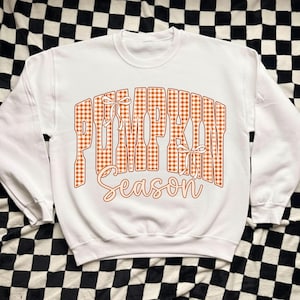 May include: A white crewneck sweatshirt featuring the text PUMPKIN SEASON in a large, orange and white gingham-patterned block font. The word PUMPKIN includes two small white line-drawn bows, and the word Season is in a cursive script.