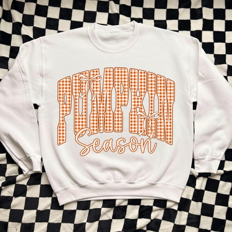 May include: A white crewneck sweatshirt featuring the text PUMPKIN SEASON in a large, orange and white gingham-patterned block font. The word PUMPKIN includes two small white line-drawn bows, and the word Season is in a cursive script.