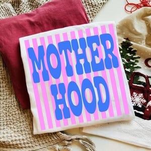 May include: A white t-shirt featuring vertical pink stripes and the text MOTHER HOOD printed in large, bold blue letters. The shirt is folded and placed on a surface with a maroon fabric and a cream-colored textile with a red truck graphic.