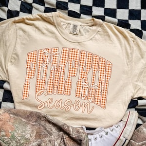May include: A beige short-sleeved t-shirt featuring the text PUMPKIN SEASON in a large orange and white gingham pattern. The word PUMPKIN is written in a blocky font with two white line-art bows, while the word Season is in a cursive script.