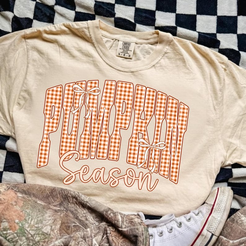 May include: A beige short-sleeved t-shirt featuring the text PUMPKIN SEASON in a large orange and white gingham pattern. The word PUMPKIN is written in a blocky font with two white line-art bows, while the word Season is in a cursive script.