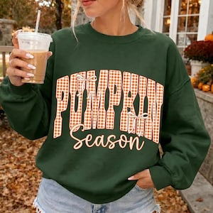 May include: A dark green long-sleeve sweatshirt featuring the text "PUMPKIN Season" in a large, orange and white gingham-patterned font. The word "PUMPKIN" is arched and decorated with two small white bows, while "Season" is written in cursive.