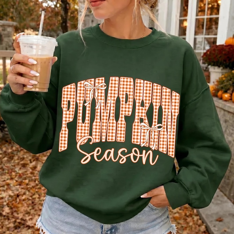 May include: A dark green long-sleeve sweatshirt featuring the text "PUMPKIN Season" in a large, orange and white gingham-patterned font. The word "PUMPKIN" is arched and decorated with two small white bows, while "Season" is written in cursive.