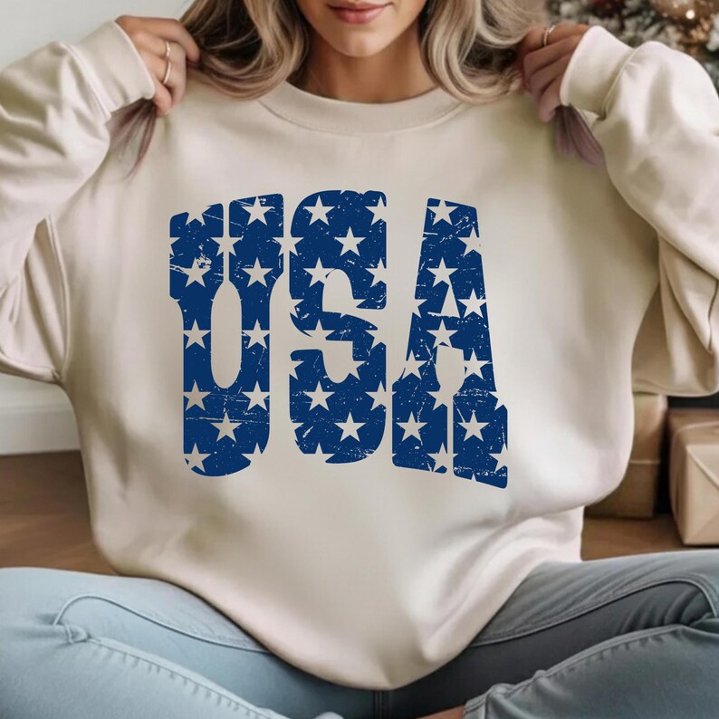 May include: Cream-colored sweatshirt with the word "USA" in distressed blue lettering filled with white stars. The sweatshirt is a neutral color, and the graphic has a vintage, patriotic feel.