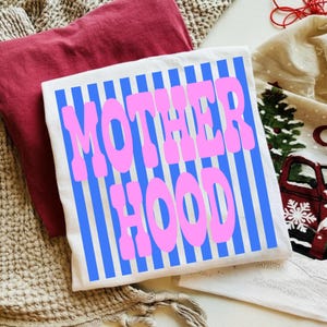 May include: A white t-shirt featuring a vertical blue striped pattern and the text MOTHER HOOD printed in large, bold pink letters across the center. The shirt is folded and displayed alongside a solid maroon fabric and a cream-colored textile.