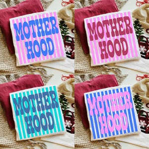 May include: Four folded t-shirts featuring a vertical striped pattern and the text MOTHER HOOD in bold, wavy lettering. The shirts are displayed in four color variations: blue on white, dark pink on white, dark blue on teal, and pink on blue.