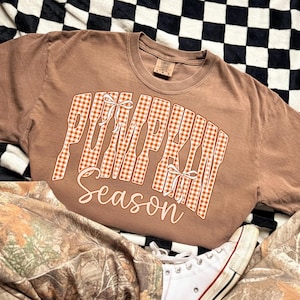 May include: A brown short-sleeve t-shirt featuring the text PUMPKIN in a white and orange gingham pattern with two small white bows, and the word season written in a cursive white font below the block letters on the front of the garment.