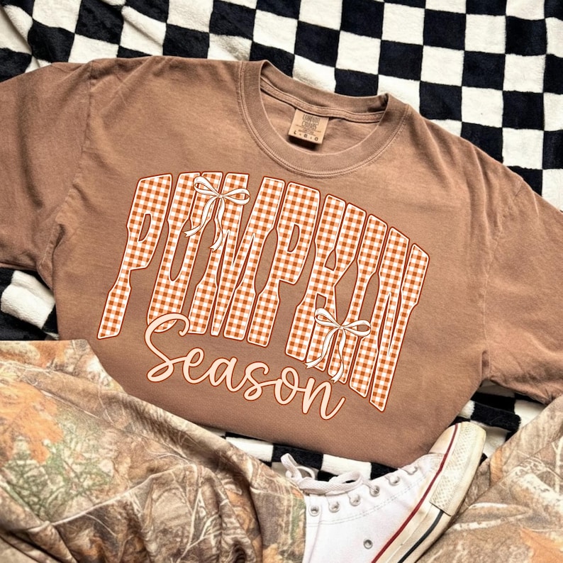 May include: A brown short-sleeve t-shirt featuring the text PUMPKIN in a white and orange gingham pattern with two small white bows, and the word season written in a cursive white font below the block letters on the front of the garment.