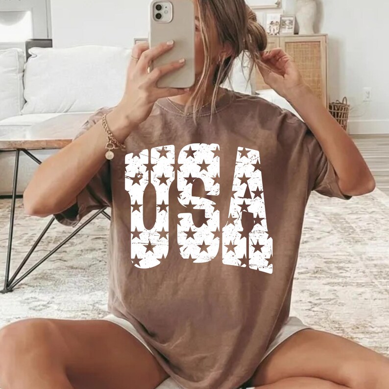 May include: A brown t-shirt with the word "USA" printed in large, distressed white letters filled with star patterns. The shirt has short sleeves and a relaxed fit. The image shows the shirt being worn.