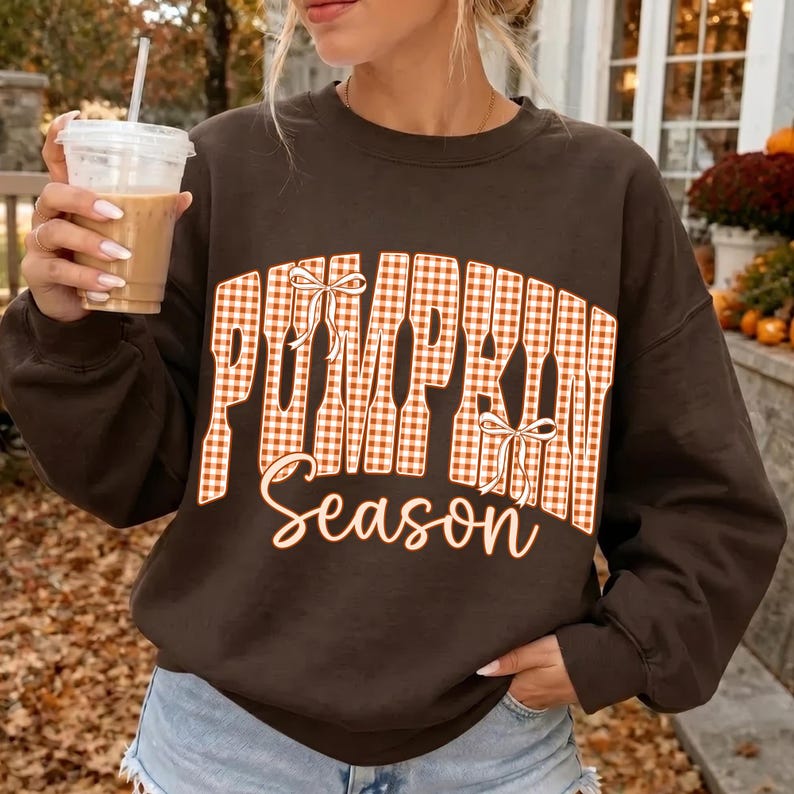 May include: A dark brown crewneck sweatshirt featuring the text "PUMPKIN Season" in a white and orange gingham pattern. The word "PUMPKIN" is arched and decorated with two white line-art bows, while "Season" is written in a cursive script font.