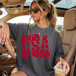 May include: Dark gray t-shirt with the word "USA" in red, star-filled letters. The shirt is worn by a person in a car, accessorized with sunglasses and gold jewelry. The person is holding a beverage.